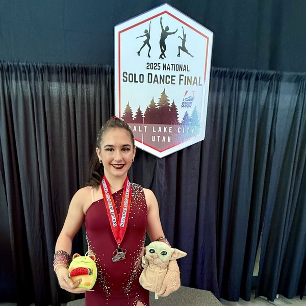 A student from The Siena School wearing a dark red ice dancing costume wears a silver medal at nationals and looks proudly at the camera. She's holding 2 stuffed animals.