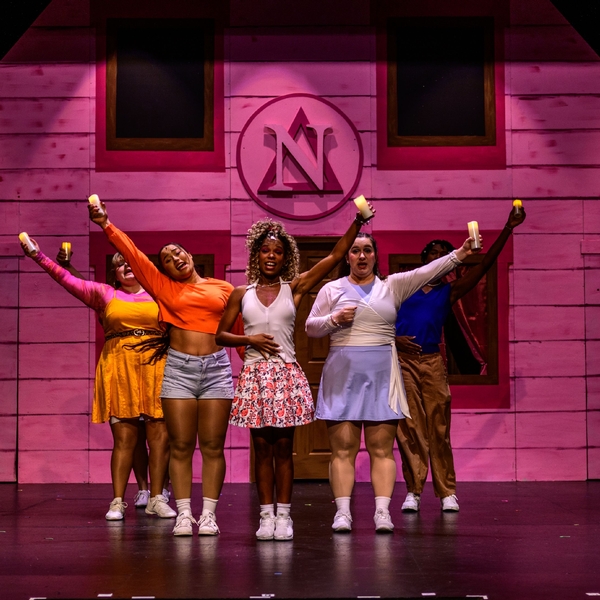 A teacher from The Siena School stands centerstage with her castmates behind her during a musical production. They are in front of a house set piece in the background, and the lighting is pink.