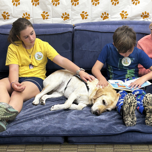 Two elementary students from The Siena School sit on a blue floor cushion as a service dog rests between them. One student is reading a book aloud.