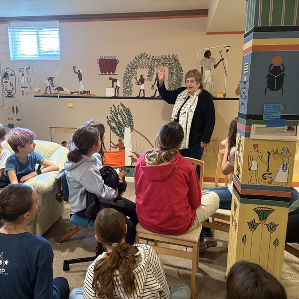 A woman stands in front of a wall adorned with her mural depicting scenes from Ancient Egypt as 6th graders from The Siena School look on with interest.