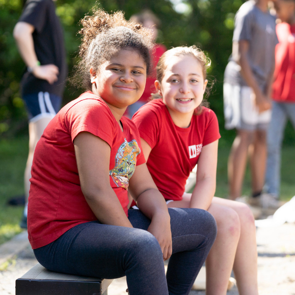 Two girls from The Siena School sit outside on a bench. The suns shines on their faces, and they smile happily.
