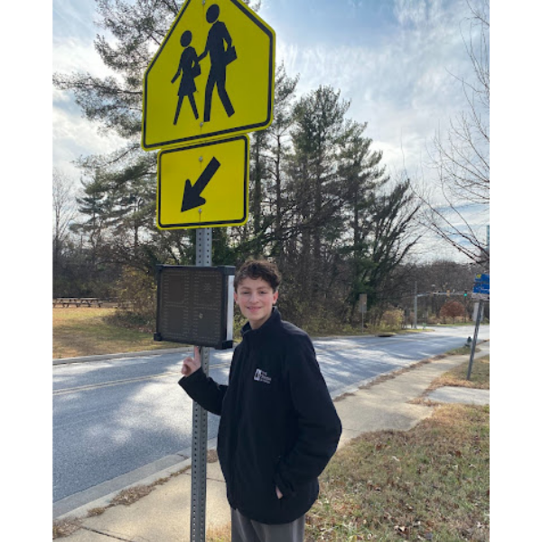A high school student stands proudly next to a street sign with a digital speed monitor. He advocated for better crosswalk safety.