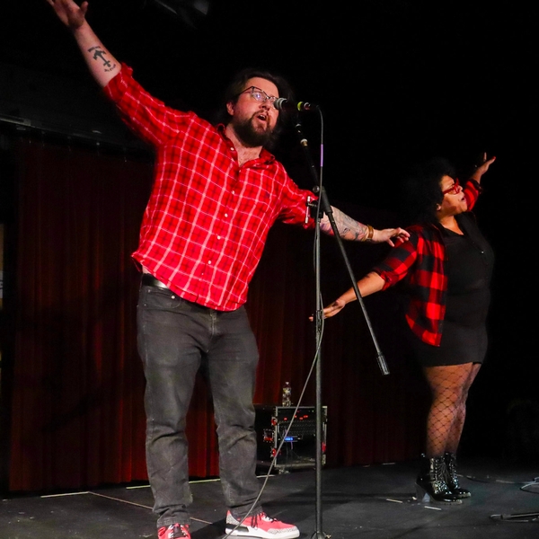 A Siena teacher in a red shirt stands on stage in front of a microphone during a spoken word poetry competition. He and his partner have their arms spread in a bird pose.