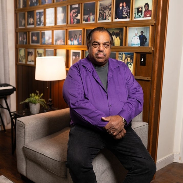 A man in a purple shirt sits on the side of an armchair looking confidently at the camera in his home office.