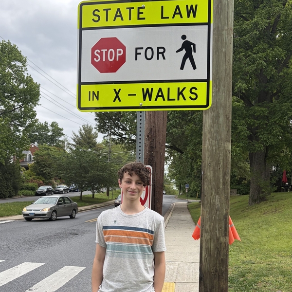 A high school student from The Siena School stands proudly under a street sign that says STATE LAW STOP FOR PEDESTRIANS. He advocated for the sign to improve crosswalk safety.