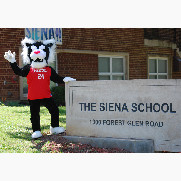The Siena School's mascot Wiley stands outdoors in front of the school sign and waves in welcome.