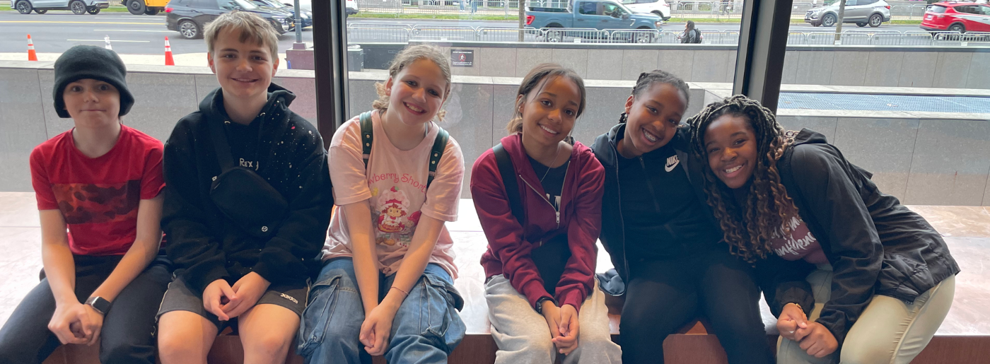 A group of 6 students sit on a bench and smile at the camera while on a field trip.