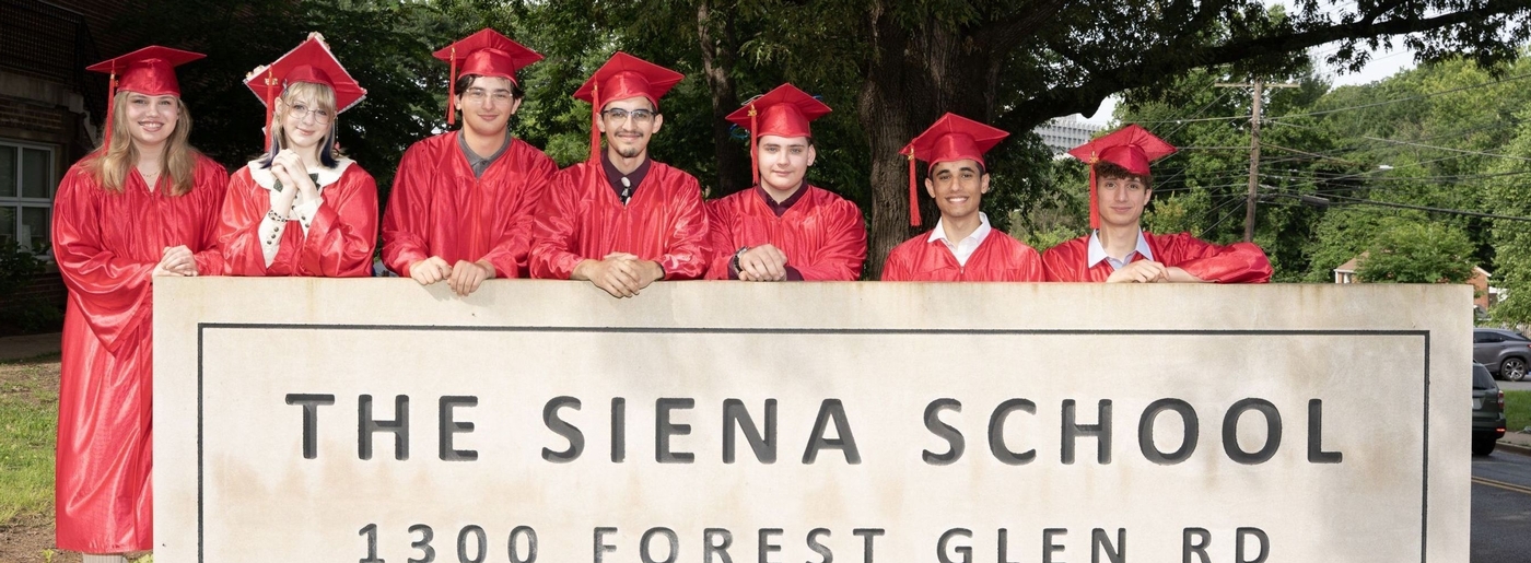 Students wearing their red caps and gowns standing near The Siena School sign on Graduation Day.