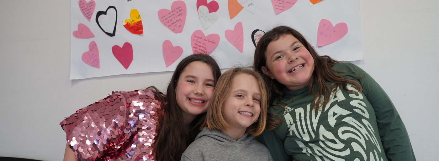 Three girls look at the camera and smile; there are paper hearts on a poster in the background.