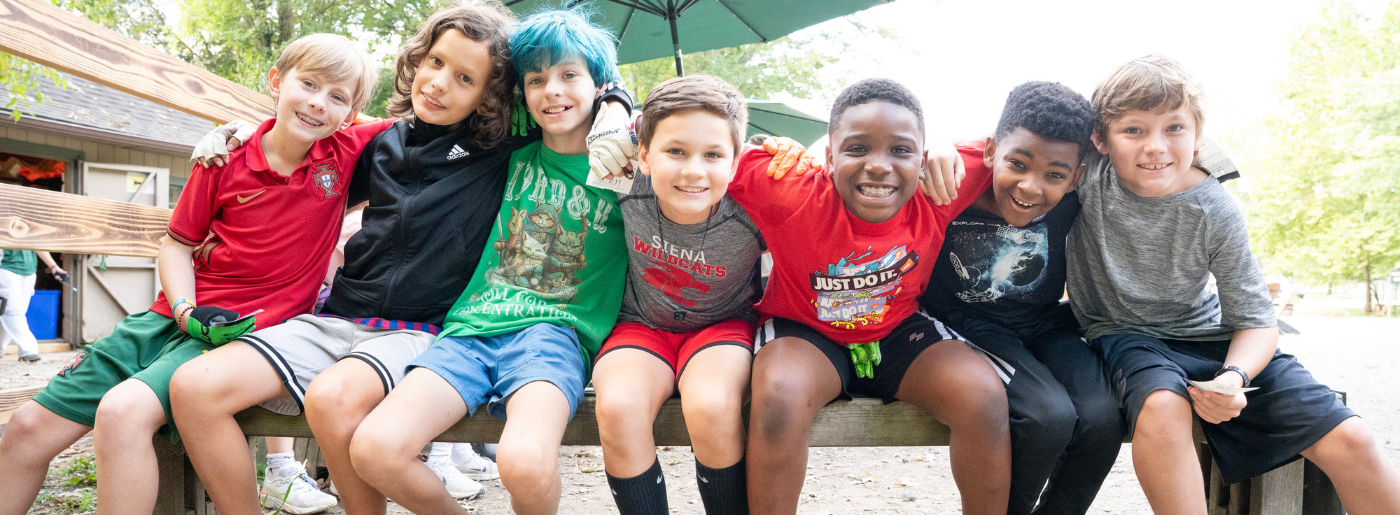 Seven boys sit on a bench outside with their arms around each other. They are very happy.
