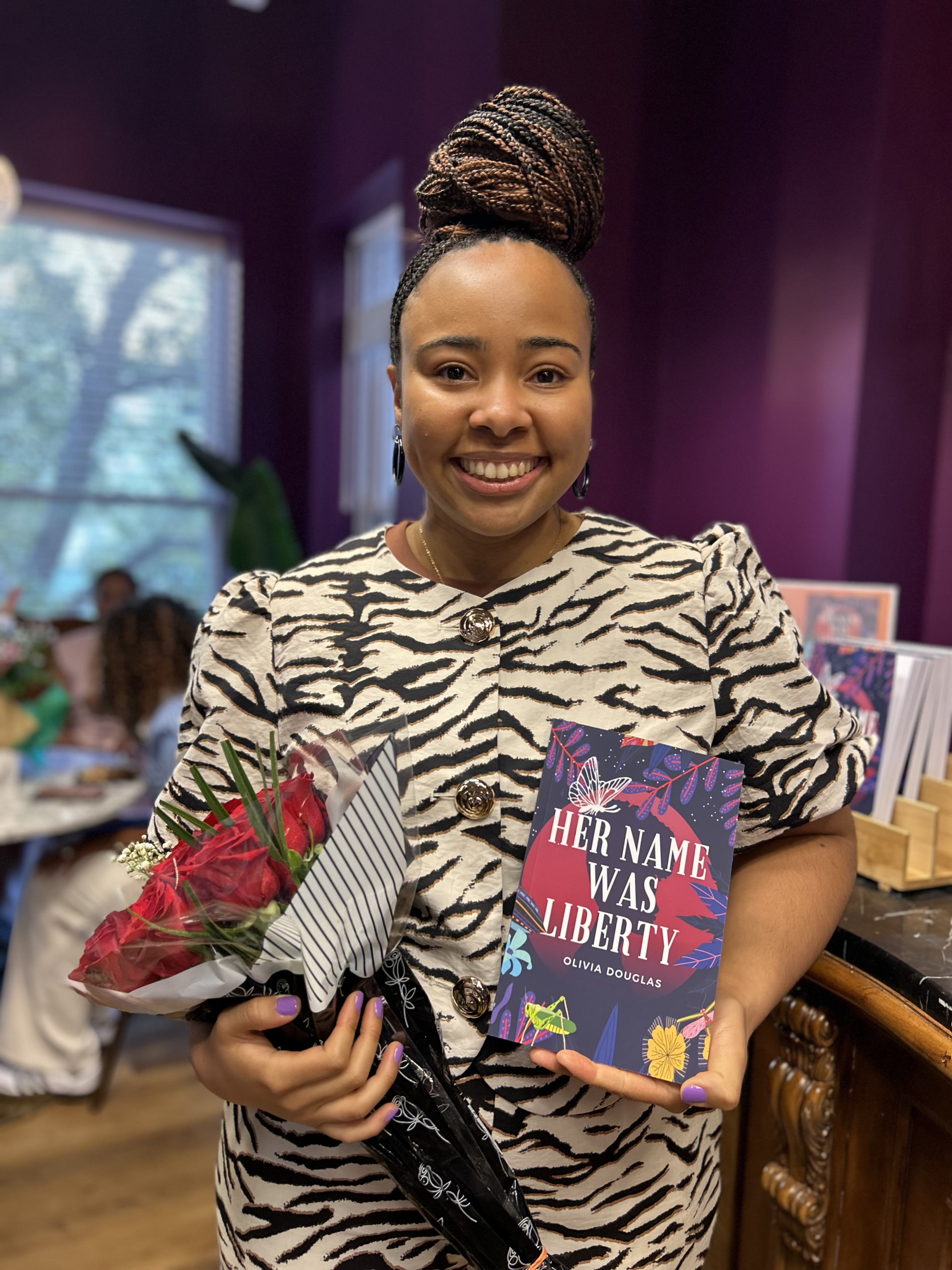 A woman smiling and holding a bouquet of red flowers and a book that she wrote.