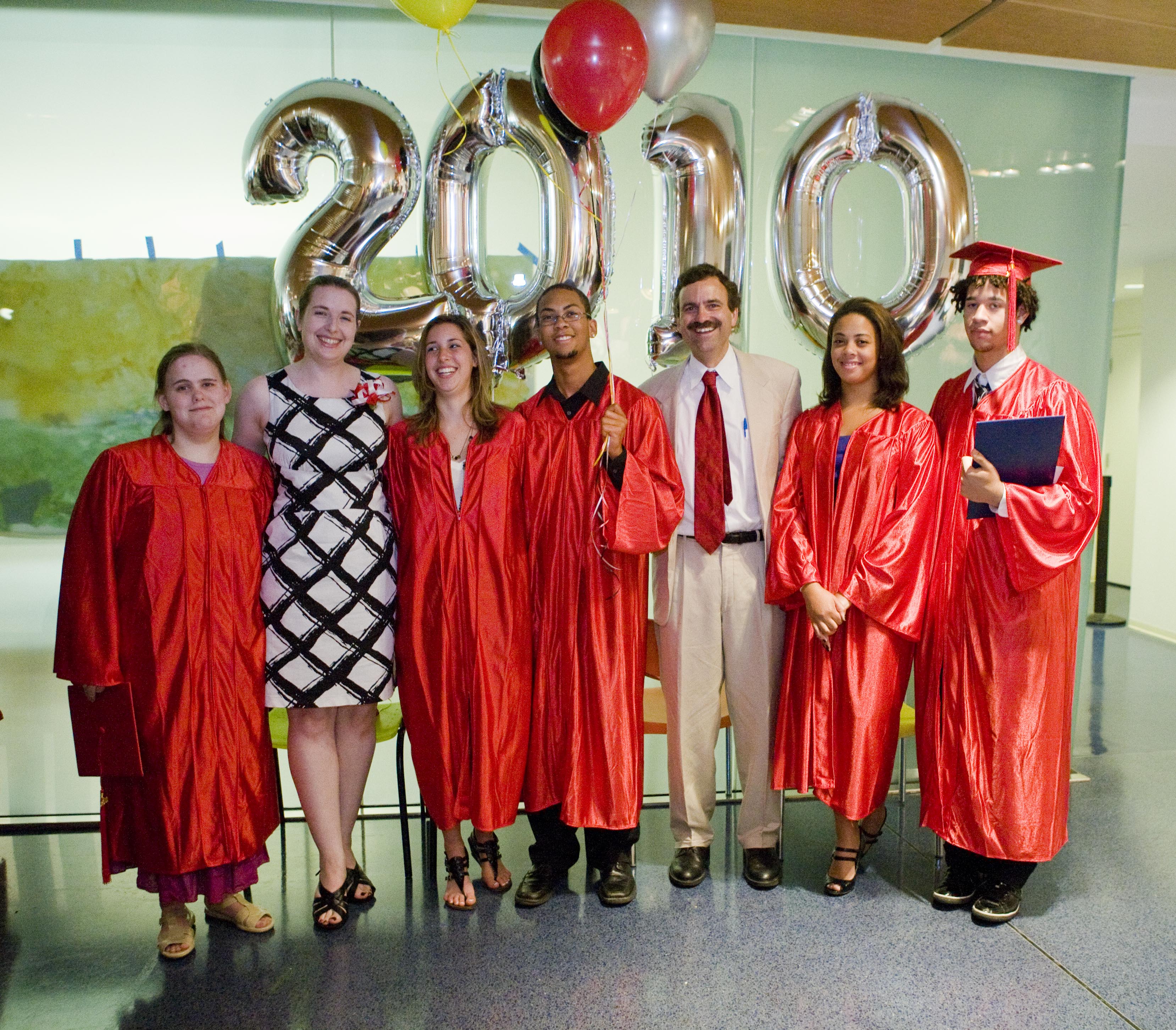 Five high school graduates in red caps and gowns joined by two nicely dressed adults on graduation day. Shiny balloons spelling 2010 are behind them.