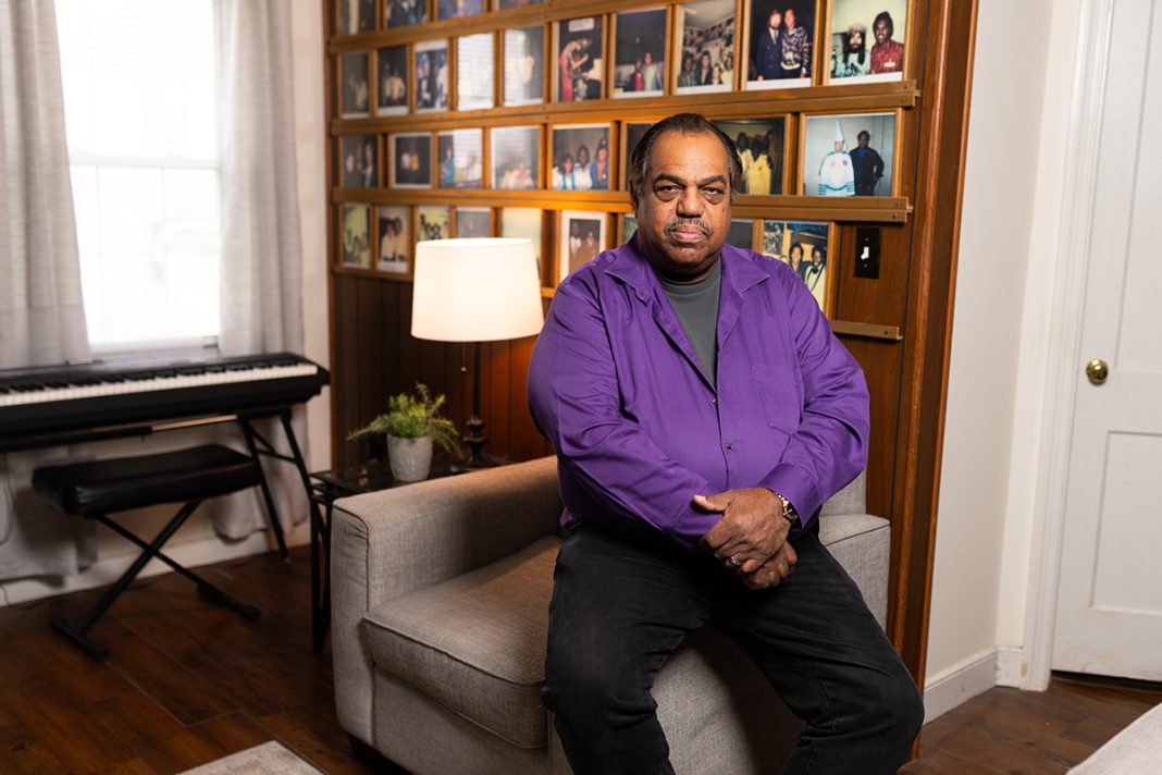 A Black man sits proudly and confidently on the arm of a chair and looks at the camera. 