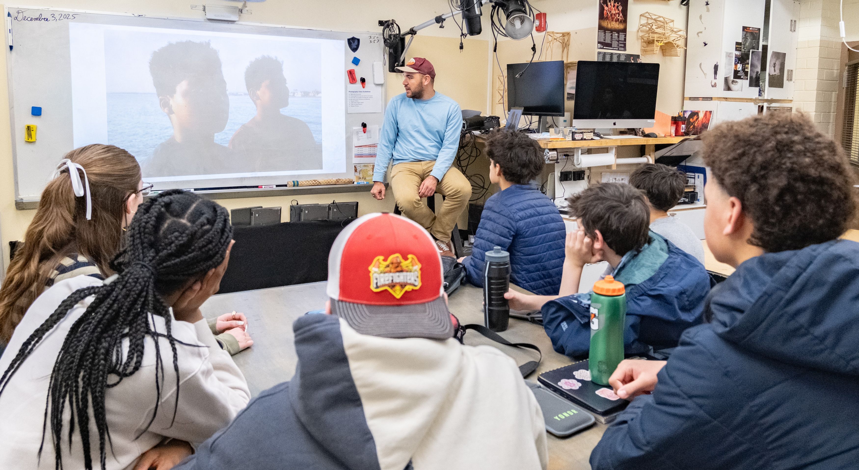 Students sit at a table and listen to a guest speaker showing his photography.