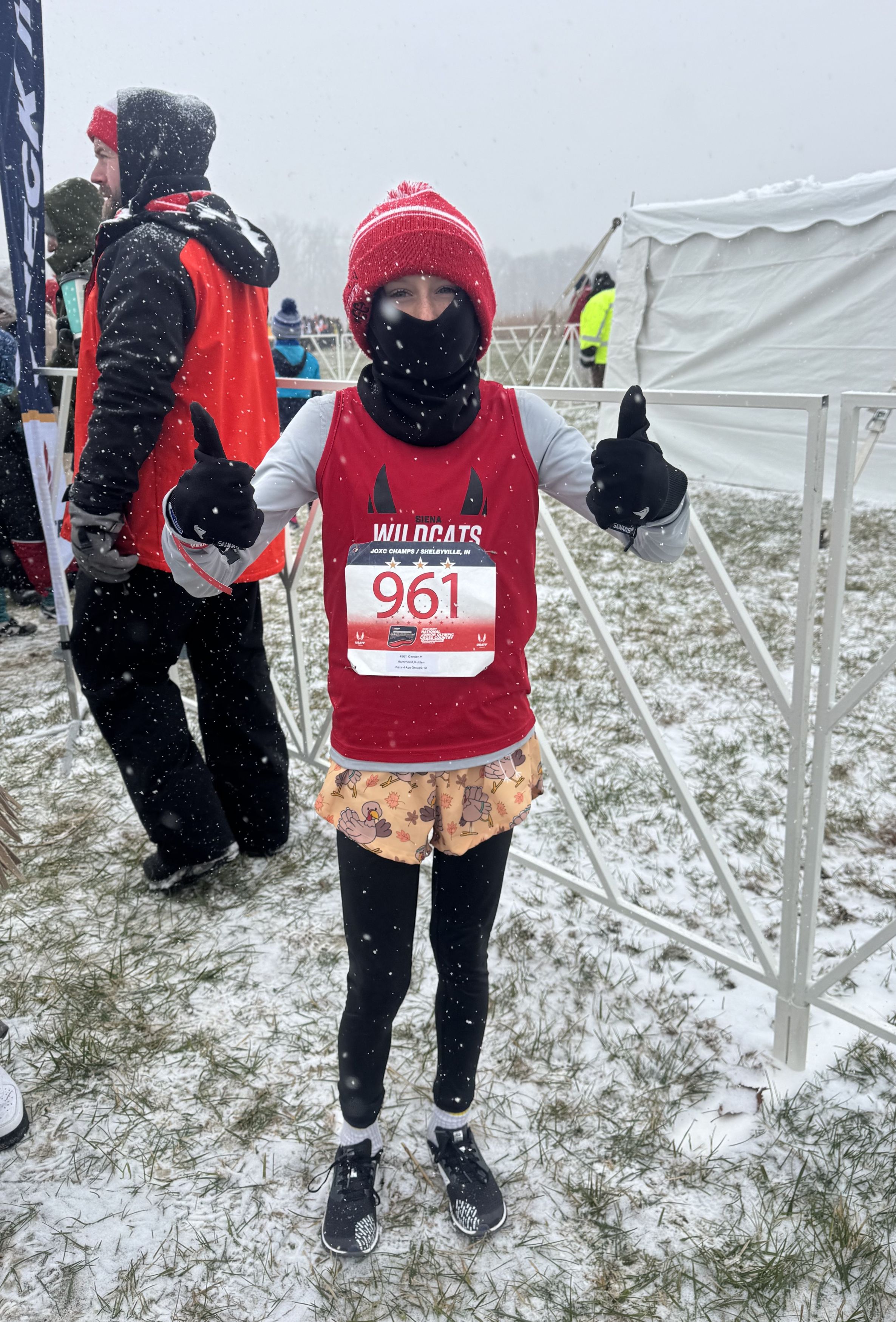 A student in a red hat and race vest gives a thumbs up while standing in the snow.