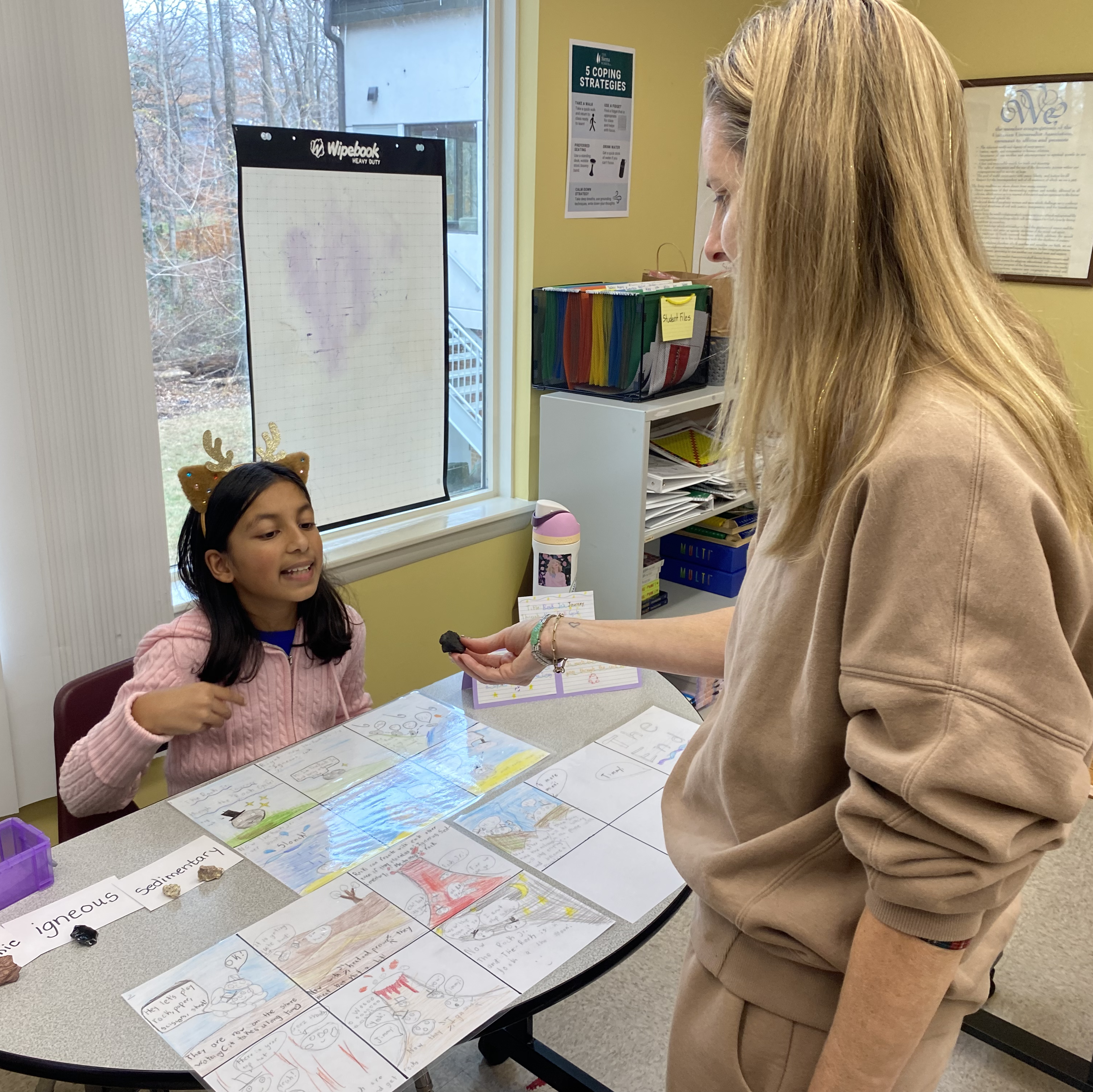 A student presents a science project at a table while a parent listens and holds a rock sample in a classroom.