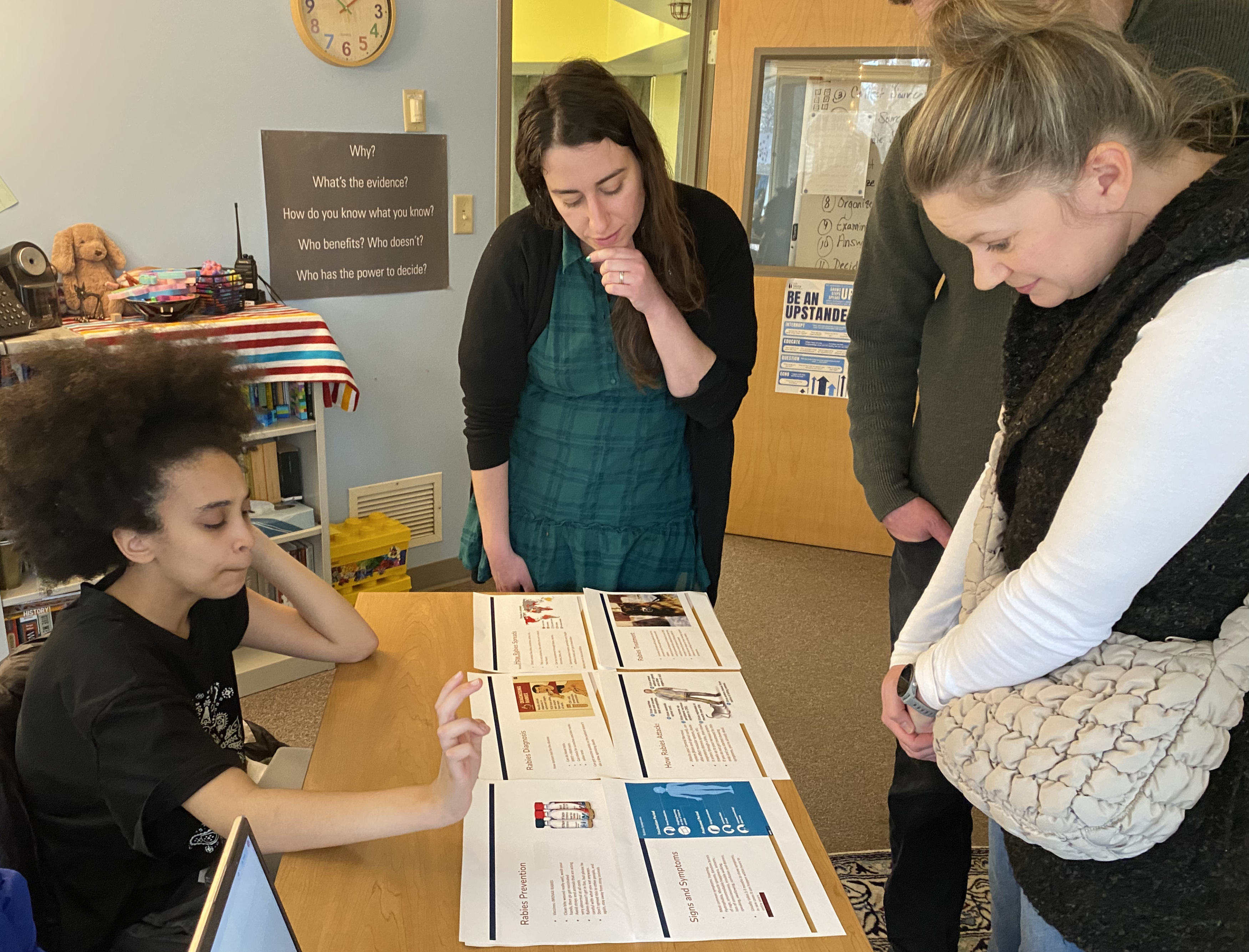 A student sits at a table with a teacher and parents standing and looking at a science project overview. 