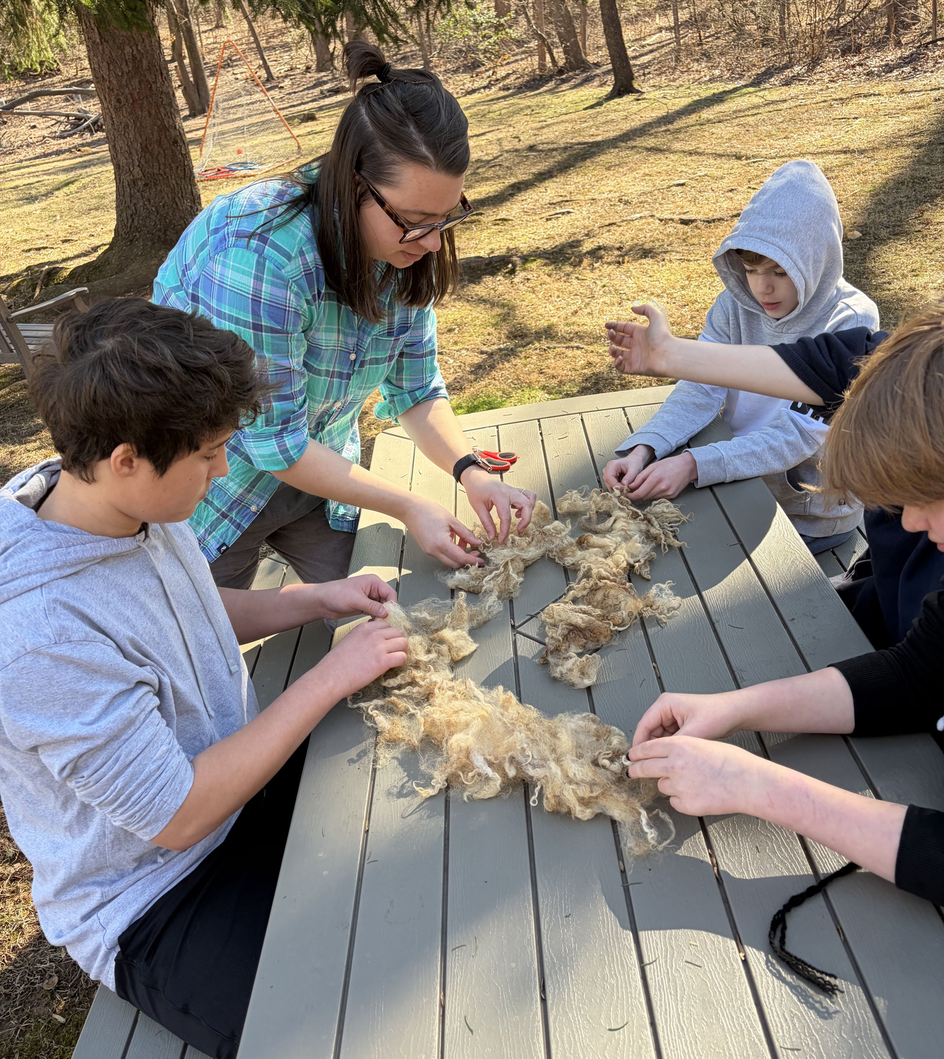 Four students and their teacher sit outdoors at a picnic table and skirt wool by hand.