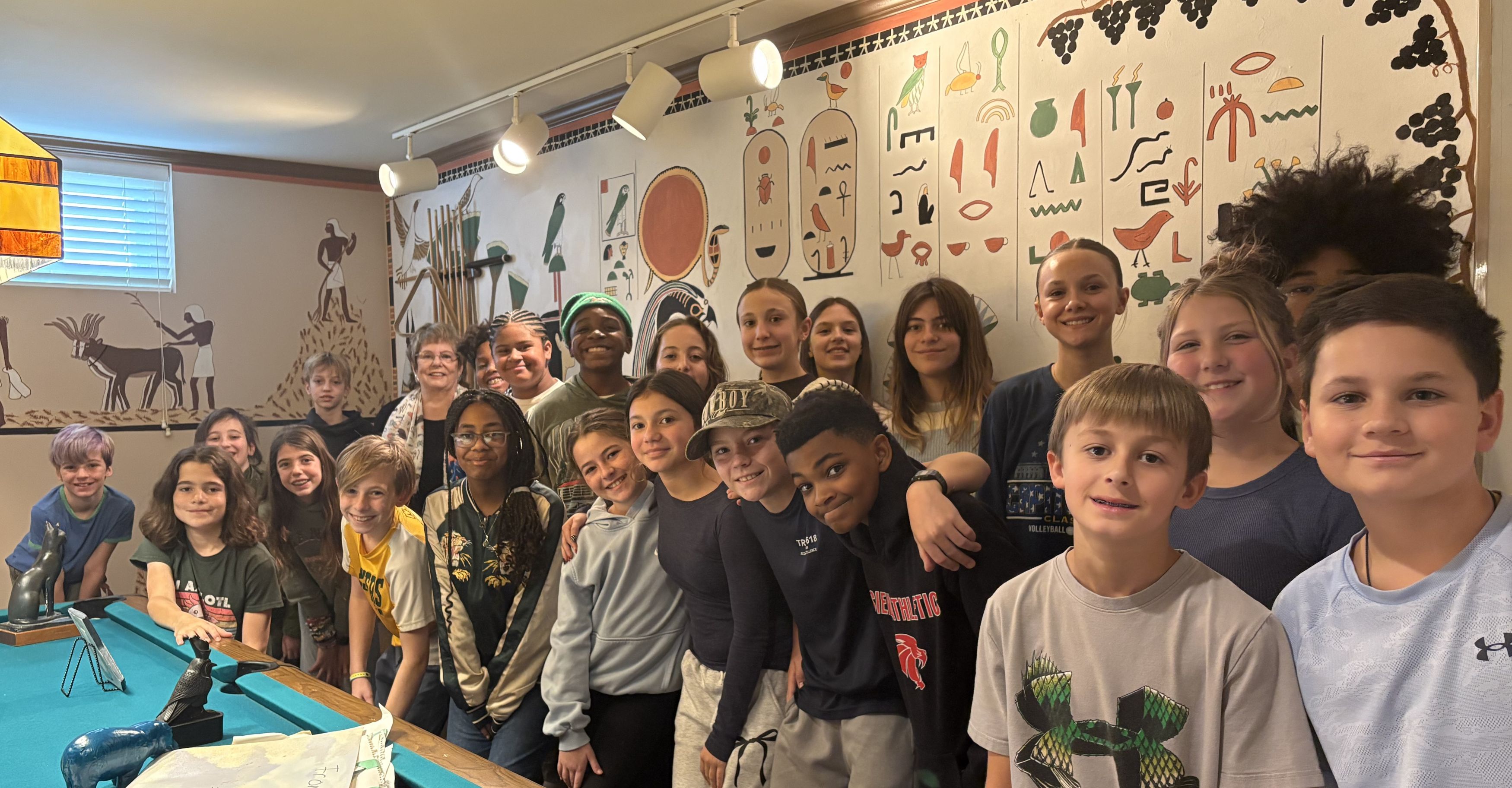 A group of middle school boys and girls stand in front of a wall mural of an Ancient Egyptian scene. They are very happy.