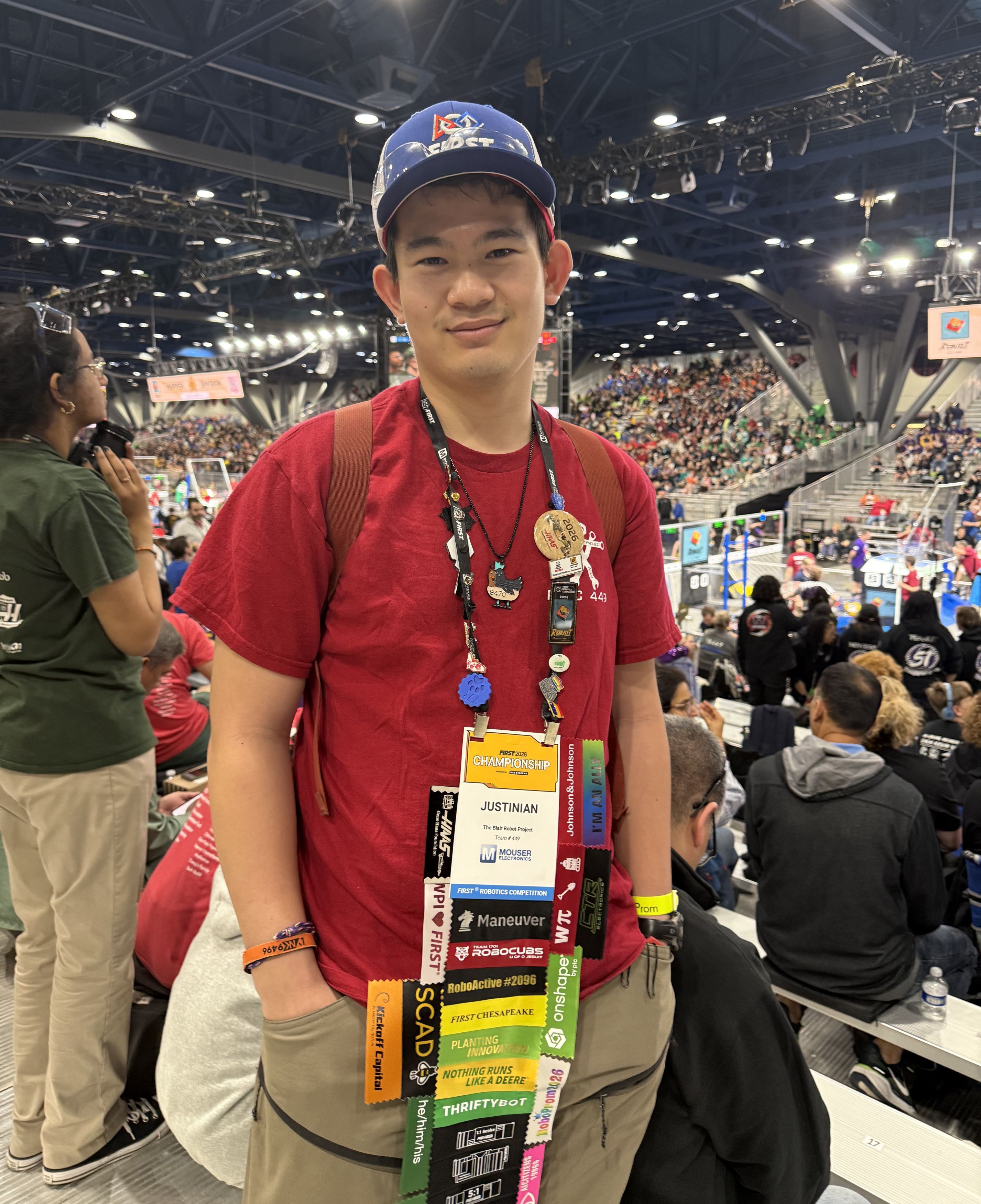 A high school boy looks proudly at the camera during a robotics competition. A large crowd is behind him.