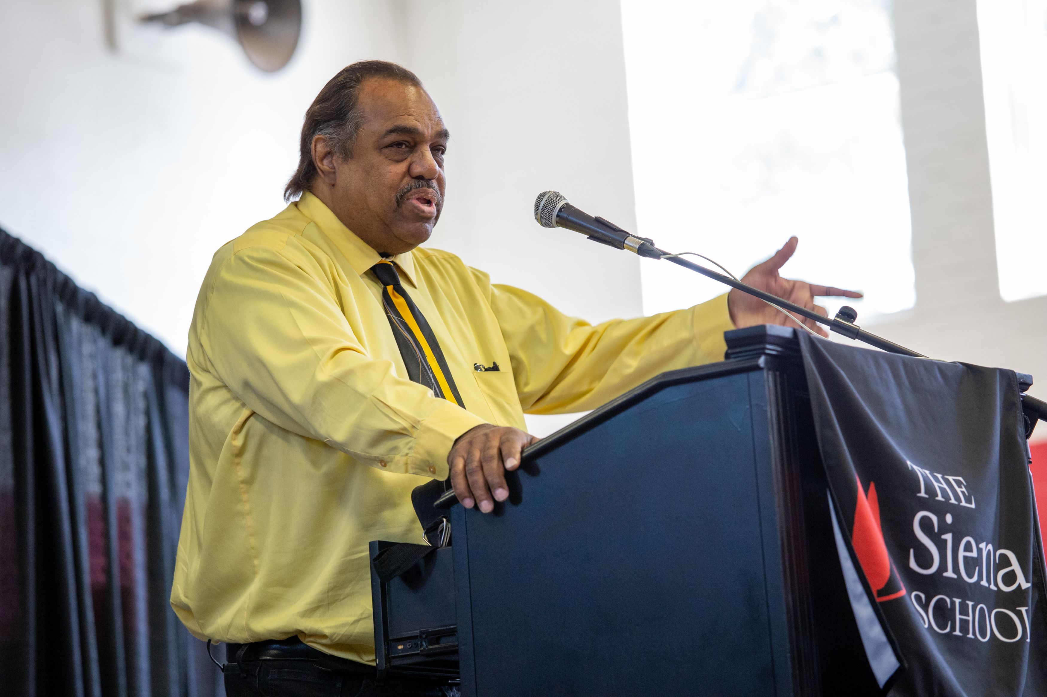 A man in a yellow shirt and striped tie stands at a lectern to deliver a commencement address.