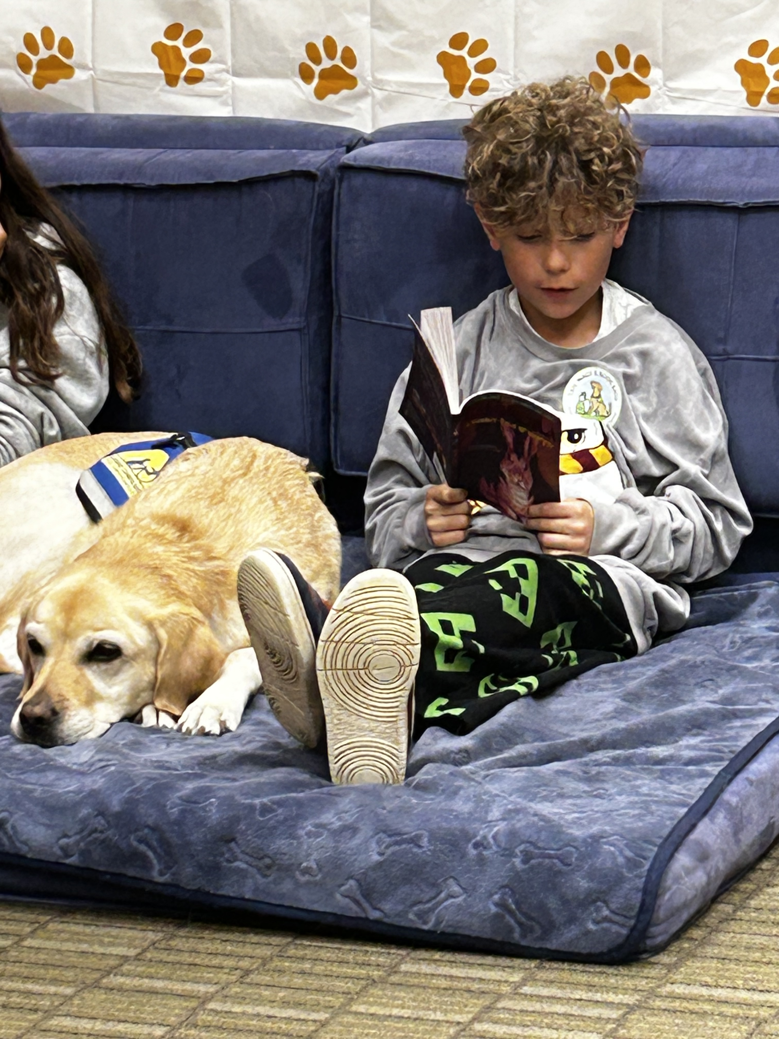 A student sits on a blue couch with an open book reading to a dog, who lazes comfortably.
