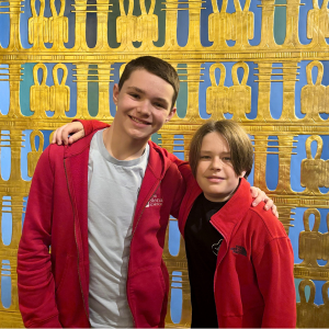 Two boys in red jackets smile at the camera in front of a gold museum display.