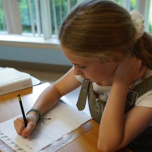 A girl sits at a desk and writes on a piece of paper. She is very focused.