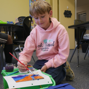 A boy in a pink hoodie kneels on the floor of a classroom and paints watercolor.