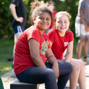 Two girls in bright red shirts smile at the camera while outside on a sunny day.