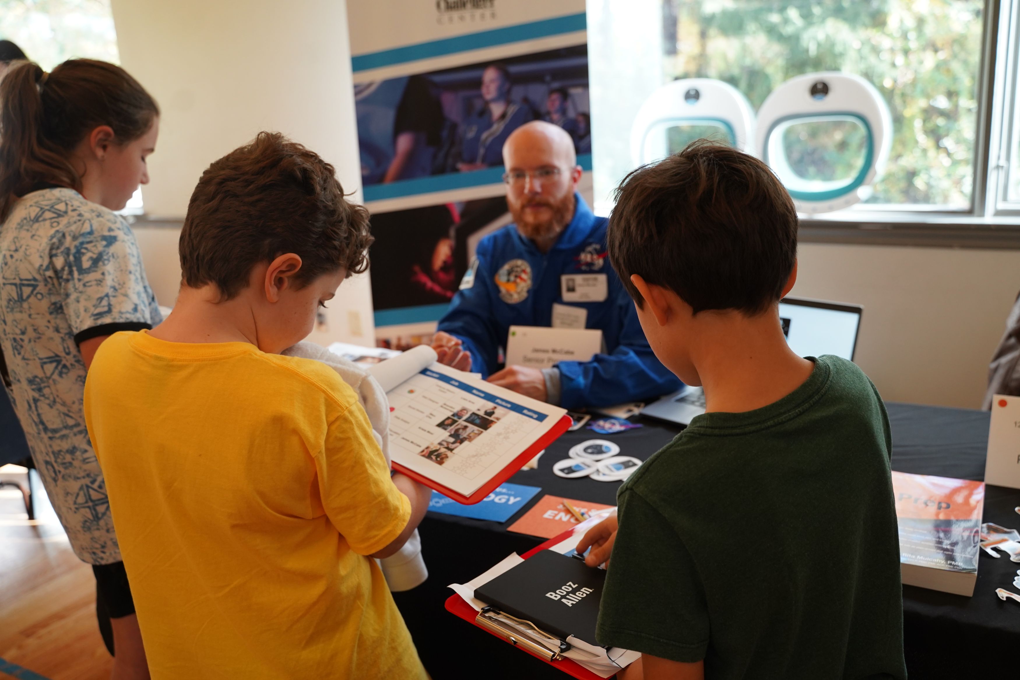Children talk with an educator in a blue NASA jumpsuit at an informational booth, holding clipboards and materials about space and STEM education..