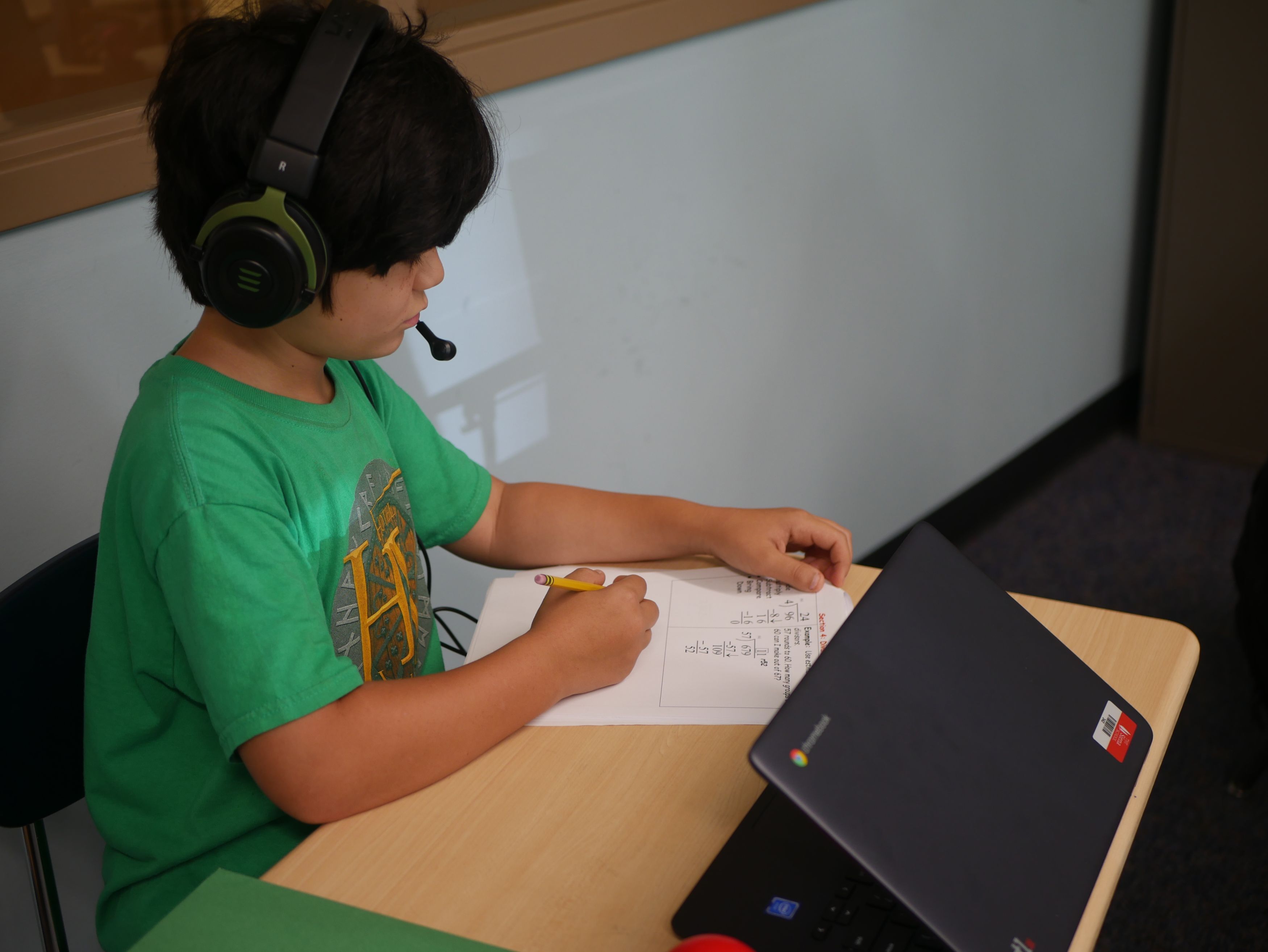 A student in a green shirt works at a desk with headphones and a laptop computer.
